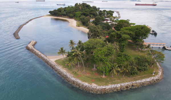 Aerial view of a tropical island, with sandy beaches, lush green trees, and a stone breakwater. Ships are visible in the distance.